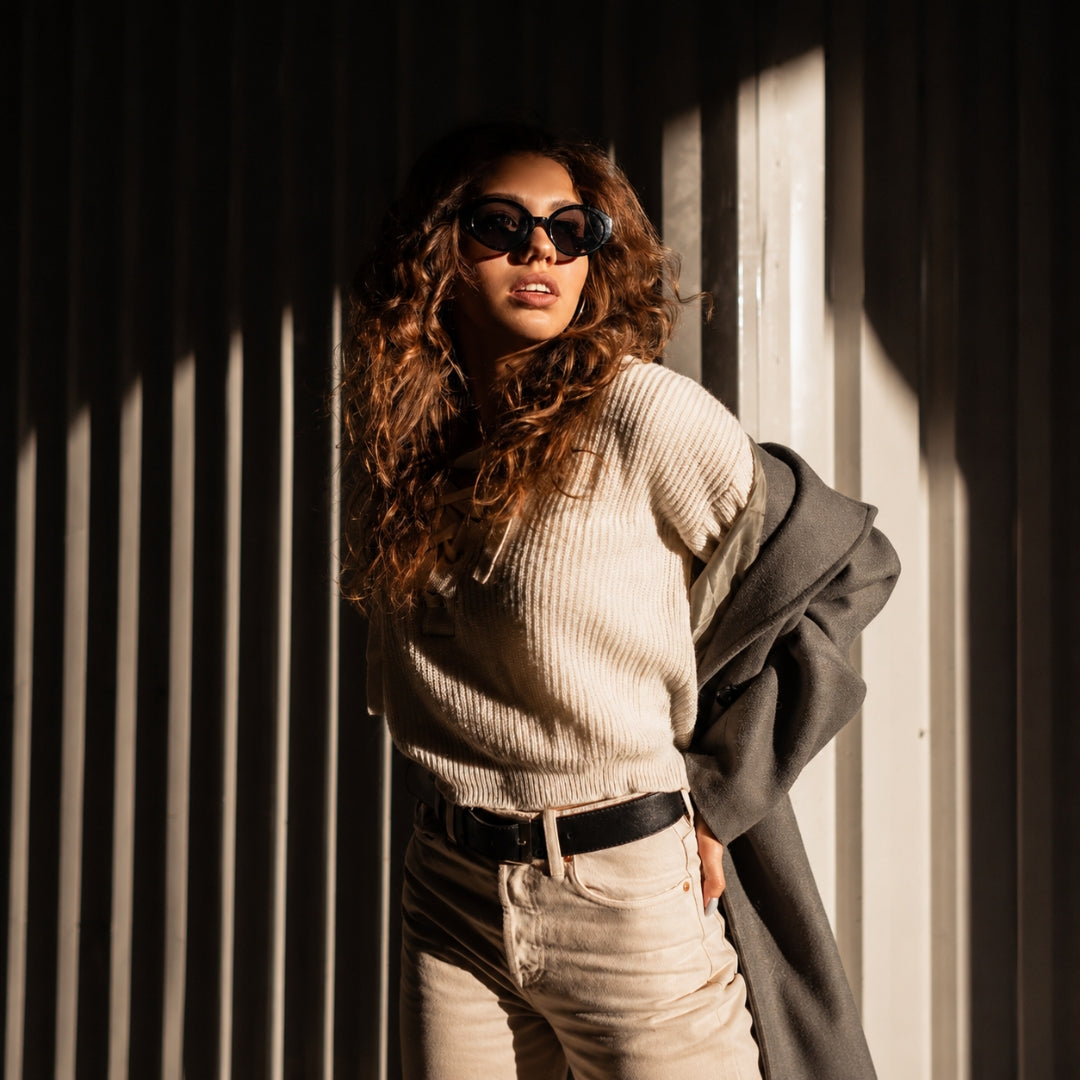 A woman with long curly brown hair wearing black sunglasses, a beige ribbed knit sweater, beige jeans, and a dark gray coat draped over her arm, posing against a corrugated metal wall with dramatic diagonal shadows.