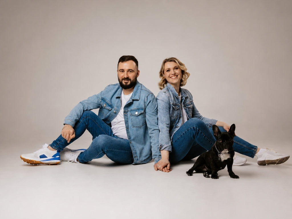 A man and a woman sitting back-to-back on a studio floor with a small black dog in front of them. Both are wearing light blue denim jackets and blue jeans, with white sneakers.