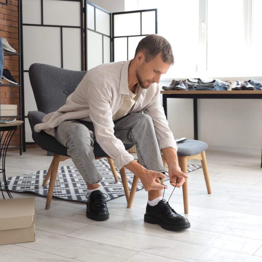 A man sitting in a gray armchair in a modern room, tying the laces of a black leather dress shoe. He is wearing a light beige button-up shirt, light gray jeans, and white socks.