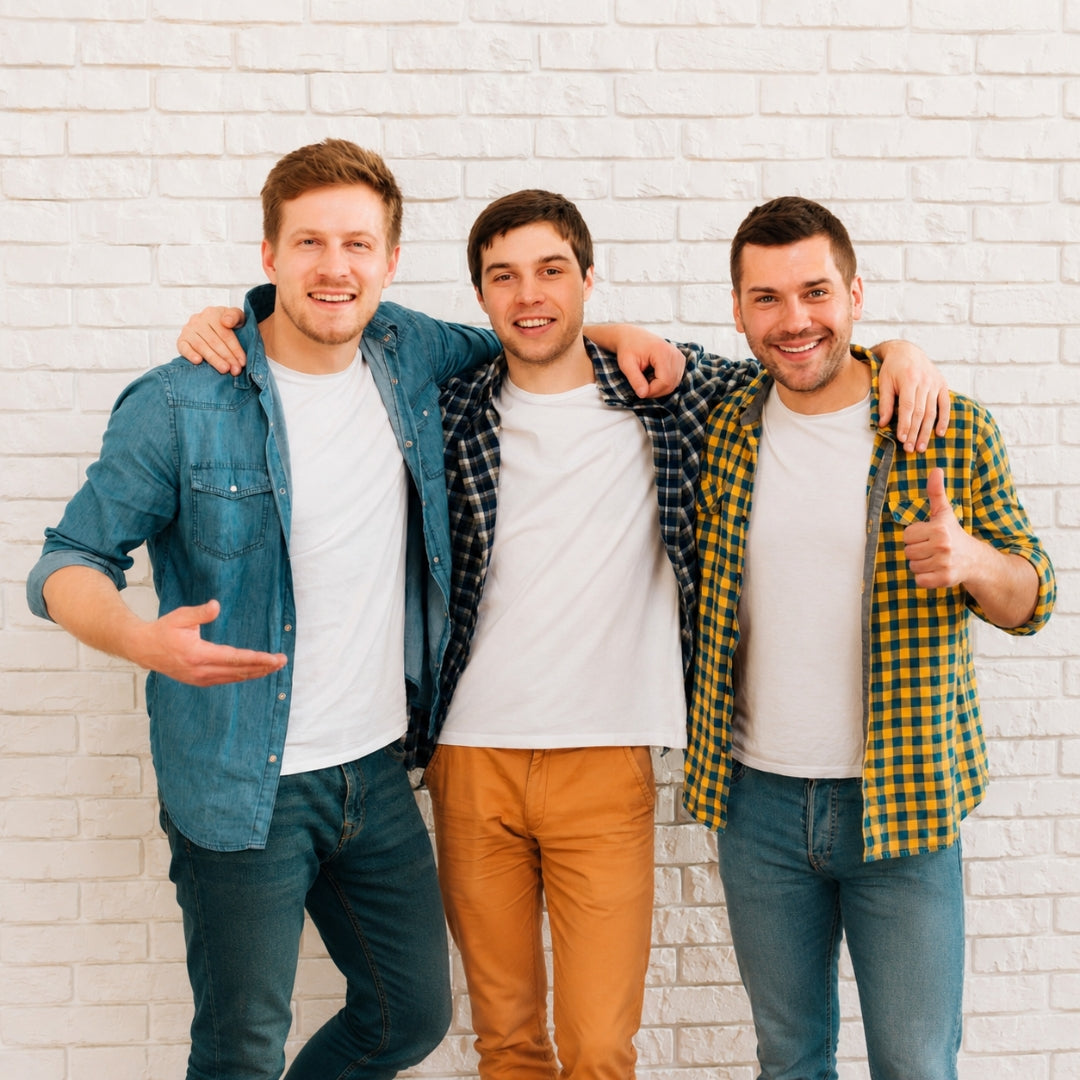 Three men standing together with their arms around each other, smiling and posing in front of a white brick wall. They are wearing casual button-up shirts and jeans in blue, yellow, and black plaid.