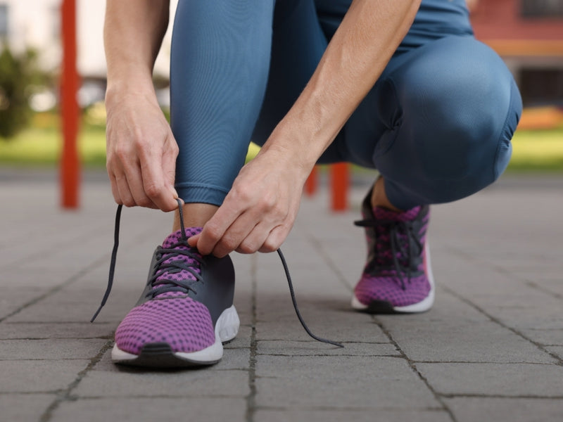 A close-up lifestyle shot of a person crouching outdoors and tying the laces of their purple and gray running shoes. The person is wearing blue-gray leggings, and the background shows a blurred paved path with red poles.