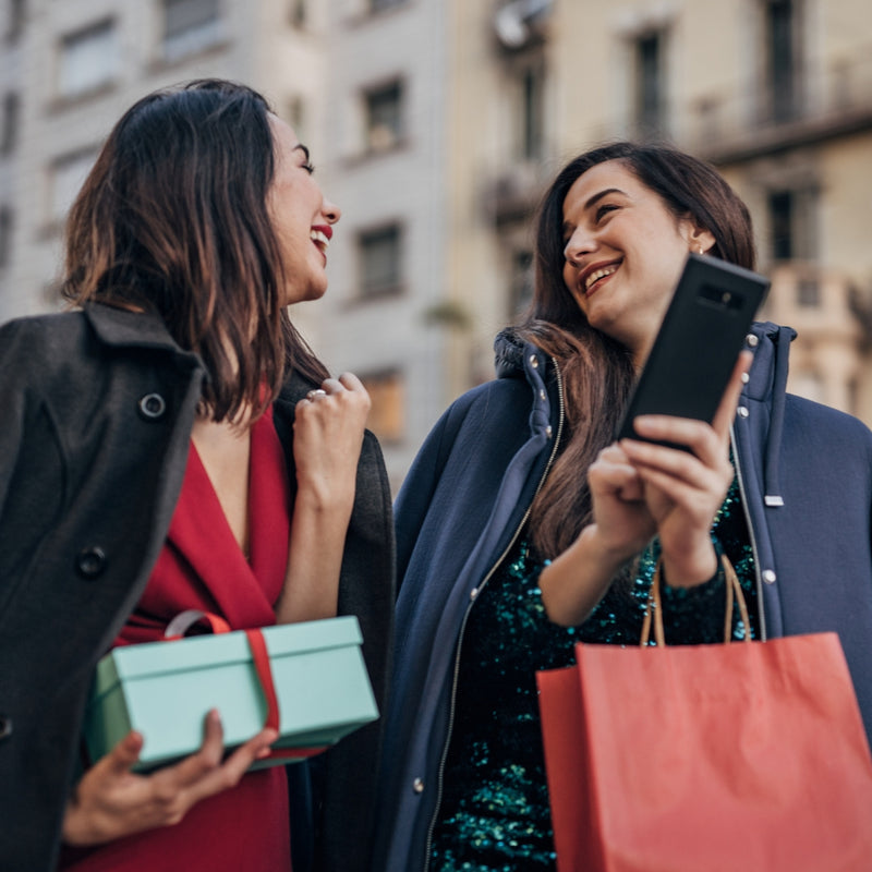 Two women smiling and laughing outdoors while shopping, one holding a mint green gift box and the other holding a red shopping bag and a black smartphone.