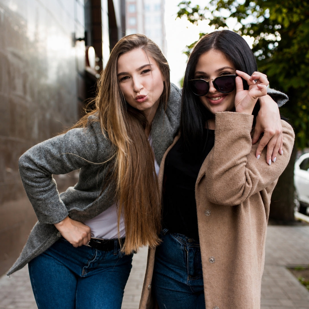 Two women posing together outdoors on a city sidewalk. The woman on the left wears a gray cardigan over a white top and blue jeans, while the woman on the right wears a camel beige cardigan over a black top and blue jeans, holding black sunglasses.